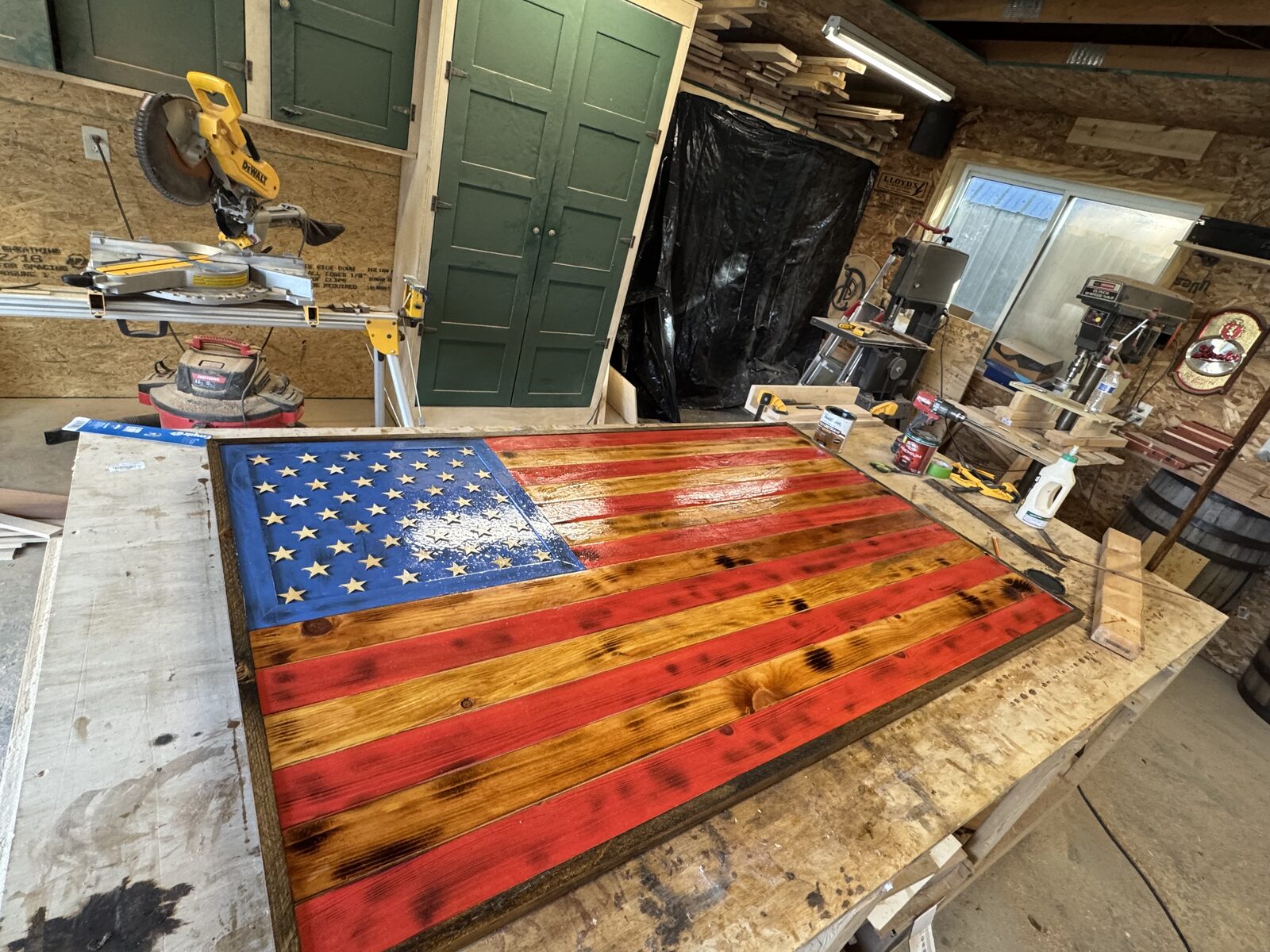 Handcrafted wooden American flag with red stained and natural torched wood stripes, a blue field of wooden stars, and a dark frame, on a workshop bench